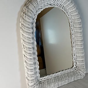Puede incluir: Un espejo de mimbre blanco arqueado. El espejo tiene forma rectangular con esquinas superiores redondeadas. El marco está hecho de mimbre tejido, pintado de blanco. El espejo está ligeramente inclinado, mostrando un reflejo.