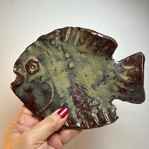 Puede incluir: Un plato artesanal de cerámica con forma de pez y superficie texturizada. El plato presenta una paleta de colores marrón, verde y beige. El pez tiene aletas detalladas y un ojo redondo. El plato está diseñado para uso decorativo o funcional.