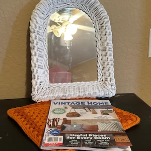 Puede incluir: Espejo blanco arqueado de mimbre con cristal rectangular. Una pila de revistas titulada "VINTAGE HOME" descansa sobre un cuadrado de ganchillo naranja. La portada de la revista presenta artículos de decoración del hogar. El espejo está colocado sobre una superficie oscura.