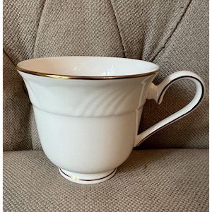 May include: White teacup with a gold rim and handle trim. The cup has a classic design with a slightly flared top and a decorative embossed pattern. The handle is curved and elegant, and the cup sits on a small foot.