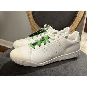 May include: A pair of white low-top sneakers featuring bright green laces and three rows of perforated stripes on the side panels. The shoes have a smooth white upper, a reinforced toe cap, and a white rubber sole, with a tag attached to the laces.