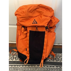 Puede incluir: Una mochila de color naranja brillante con un panel central negro y una base negra. La solapa superior muestra un logotipo triangular negro con las letras ACG. La bolsa incluye correas ajustables, bolsillos laterales y un cierre de cordón.