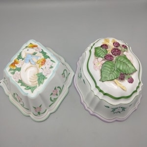 May include: Two white ceramic kitchen molds with scalloped edges. One mold is square-shaped featuring a white rabbit with a blue bow and butterflies. The other is oval-shaped, decorated with a raised cluster of green leaves and purple blackberries.