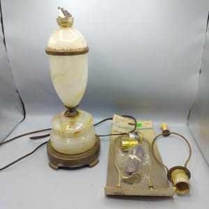 May include: A table lamp with a cream-colored stone body and a brass base, featuring a tall, urn-shaped top section. Beside the lamp are loose brass lamp parts, including a harp, a socket, and a package labeled 3-Way Turn Knob Socket.
