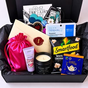 Puede incluir: Una caja de regalo negra que contiene una bolsa de satén rosa con cordón, un tubo de crema de manos con colágeno Korean Garden, una vela aromática Teakwood and Tobacco en frasco negro, palomitas Smartfood y una bolsita de té Earl Grey.