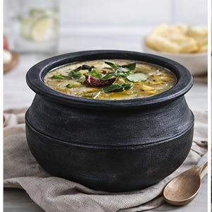 Puede incluir: Una olla redonda negra y texturizada llena de sopa, adornada con hierbas verdes e ingredientes rojos. Una cuchara de madera descansa sobre un paño beige junto a la olla. Un vaso de agua y un cuenco de pan están en el fondo.