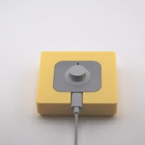 K&ouml;nnte beinhalten: Ein gelbes quadratisches Ger&auml;t mit einem grauen Zentrum und einem Drehknopf. Ein graues Kabel erstreckt sich von unten. Das Ger&auml;t befindet sich auf wei&szlig;em Hintergrund. Das Ger&auml;t scheint ein elektronisches Bauteil zu sein.