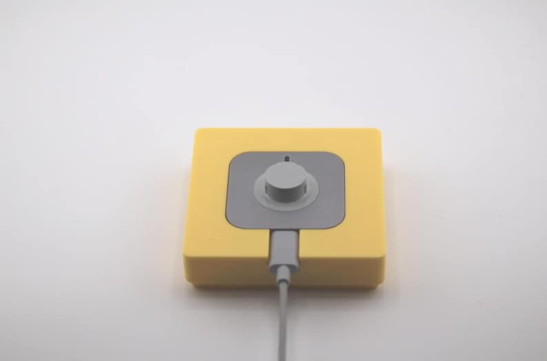 K&ouml;nnte beinhalten: Ein gelbes quadratisches Ger&auml;t mit einem grauen Zentrum und einem Drehknopf. Ein graues Kabel erstreckt sich von unten. Das Ger&auml;t befindet sich auf wei&szlig;em Hintergrund. Das Ger&auml;t scheint ein elektronisches Bauteil zu sein.