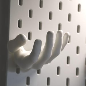 Könnte beinhalten: Ein weißer, handförmiger Wandhaken, montiert auf einer weißen Lochwand. Der Haken ist zum Aufhängen von Gegenständen konzipiert und verleiht jedem Raum eine einzigartige, künstlerische Note. Die Lochwand hat gleichmäßig verteilte, ovale Löcher.