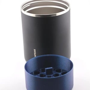 Puede incluir: Un vaso negro con borde plateado y la palabra "APEAK" impresa en el lateral. Un inserto azul cil&iacute;ndrico con m&uacute;ltiples agujeros se encuentra delante del vaso. El vaso est&aacute; probablemente dise&ntilde;ado para contener bebidas.
