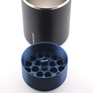 May include: A black insulated tumbler with a silver rim and the word "HYDRAPEAK" printed on the side. A dark blue, circular insert with multiple small holes sits below the tumbler.