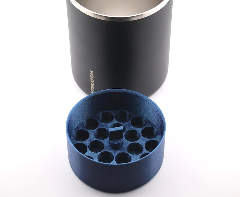 Puede incluir: Un vaso t&eacute;rmico negro con borde plateado y la palabra "HYDRAPEAK" impresa en el lateral. Un inserto circular azul oscuro con m&uacute;ltiples agujeros peque&ntilde;os se encuentra debajo.