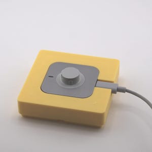 K&ouml;nnte beinhalten: Ein gelbes quadratisches Ger&auml;t mit einem grauen Mittelteil und einem kreisf&ouml;rmigen Knopf. Ein graues Kabel erstreckt sich von der Seite. Das Ger&auml;t befindet sich auf wei&szlig;em Hintergrund.