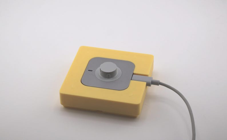 K&ouml;nnte beinhalten: Ein gelbes quadratisches Ger&auml;t mit einem grauen Mittelteil und einem kreisf&ouml;rmigen Knopf. Ein graues Kabel erstreckt sich von der Seite. Das Ger&auml;t befindet sich auf wei&szlig;em Hintergrund.