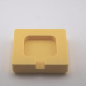 K&ouml;nnte beinhalten: Eine quadratische, gelbe Silikonform mit einem vertieften quadratischen Innenraum und einer kleinen rechteckigen Kerbe auf einer Seite. Die Form ist f&uuml;r die Herstellung oder Erstellung kleiner, quadratischer Gegenst&auml;nde konzipiert.