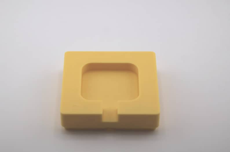 K&ouml;nnte beinhalten: Eine quadratische, gelbe Silikonform mit einem vertieften quadratischen Innenraum und einer kleinen rechteckigen Kerbe auf einer Seite. Die Form ist f&uuml;r die Herstellung oder Erstellung kleiner, quadratischer Gegenst&auml;nde konzipiert.