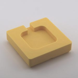 K&ouml;nnte beinhalten: Ein quadratisches, gelbes Schaumstoffobjekt mit einer vertieften Mitte und einer rechteckigen Kerbe auf einer Seite. Das Objekt ist einfarbig und liegt auf einer wei&szlig;en Oberfl&auml;che. Das Design ist einfach und geometrisch.