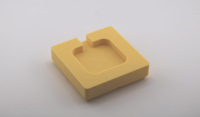 K&ouml;nnte beinhalten: Ein quadratisches, gelbes Schaumstoffobjekt mit einer vertieften Mitte und einer rechteckigen Kerbe auf einer Seite. Das Objekt ist einfarbig und liegt auf einer wei&szlig;en Oberfl&auml;che. Das Design ist einfach und geometrisch.