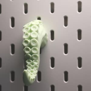 Puede incluir: Un objeto en forma de tentáculo verde claro con ventosas, montado en una pared perforada blanca. El tentáculo es de color verde pálido y tiene una superficie texturizada. La pared perforada tiene agujeros ovalados espaciados.