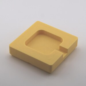 K&ouml;nnte beinhalten: Ein quadratisches, gelbes Objekt mit einem vertieften quadratischen Zentrum und einer gekerbten Seite. Das Objekt besteht aus einem festen Material und liegt auf einer wei&szlig;en Oberfl&auml;che. Das Design ist einfach und geometrisch.