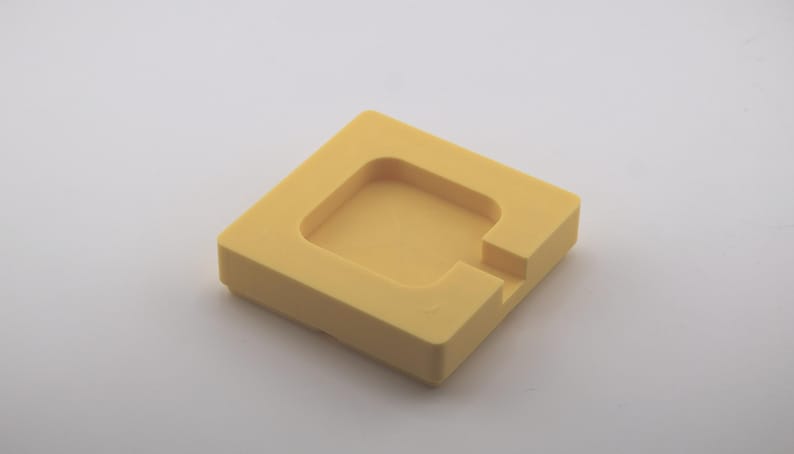K&ouml;nnte beinhalten: Ein quadratisches, gelbes Objekt mit einem vertieften quadratischen Zentrum und einer gekerbten Seite. Das Objekt besteht aus einem festen Material und liegt auf einer wei&szlig;en Oberfl&auml;che. Das Design ist einfach und geometrisch.
