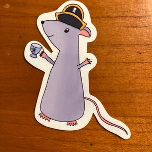 May include: A sticker featuring a cartoon rat wearing a pirate hat with a skull and crossbones. The rat is holding a goblet and has a pink tail and feet. The sticker has a white border.