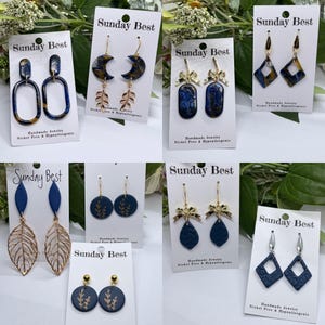 May include: A collection of handmade earrings displayed on white cards. The earrings feature various designs, including geometric shapes, leaf patterns, and bow accents. Colors include navy blue, gold, and iridescent accents. The cards are labeled "Sunday Best."