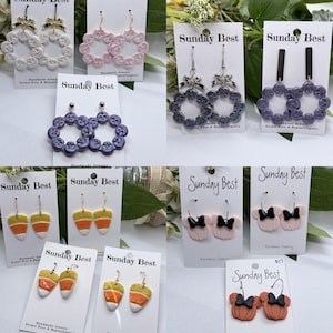 Pode incluir: Uma coleção de brincos feitos à mão, incluindo argolas em forma de botão em branco, rosa e roxo, e designs inovadores como doces de milho e brincos de laço. Cada par é exibido em um cartão com o texto "Sunday Best".