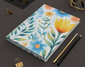 May include: A floral-patterned journal with a white background and colorful flowers in yellow, blue, and orange. The journal is open, revealing blank pages. Gold and black pencils and paper clips are nearby.