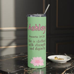 May include: A light green stainless steel tumbler with a silver straw. The tumbler features the name "Ashley" in pink script, the Bible verse Proverbs 31:25, and a pink lotus flower design.