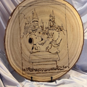 May include: Wood-burned art piece on a circular wood slice. The artwork depicts Snoopy and Harry Potter in front of Hogwarts Castle. The image is framed with a dark line and mounted on a black stand.