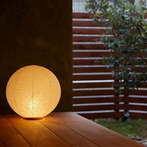 May include: A glowing, spherical paper lantern sits on a wooden surface. The warm light from within illuminates the textured paper and the surrounding area. The background features a wooden fence and greenery.