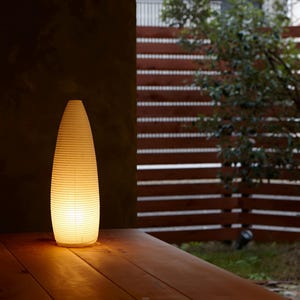 Puede incluir: Una linterna de papel blanco roto, de forma cónica, se encuentra sobre una superficie de madera. La lámpara está iluminada desde dentro, emitiendo una luz cálida. El fondo incluye una valla de madera y vegetación.