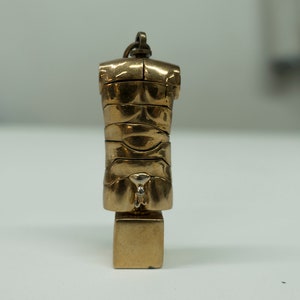 Könnte beinhalten: Ein goldfarbener Metallanhänger in Form eines menschlichen Torsos mit einem segmentierten Design. Der Anhänger ist an einer kleinen goldfarbenen Metallkette befestigt.