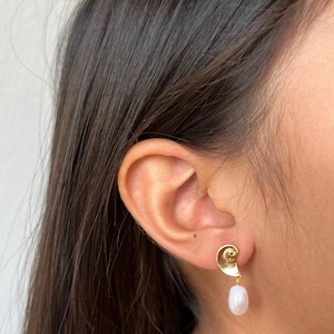 May include: A gold-coloured earring with a spiral design and a dangling white pearl. The earring is worn on a person's ear, with dark brown hair visible.