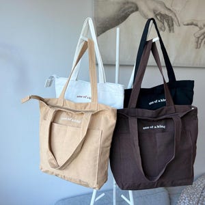 May include: Four canvas tote bags in beige, white, black, and brown, each with the text "one of a kind" printed in white. The bags have long handles and a front pocket. The bags are hanging on a white rack.
