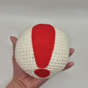 May include: A white crocheted ball with a large red exclamation point design. The exclamation point is made of felt and is centered on the ball. The ball is held in a hand.