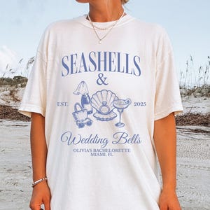 May include: White t-shirt with a blue design featuring the text "SEASHELLS & Wedding Bells" and illustrations of seashells, a cocktail, and a beach bag. The text "OLIVIA'S BACHELORETTE MIAMI, FL" is also included.