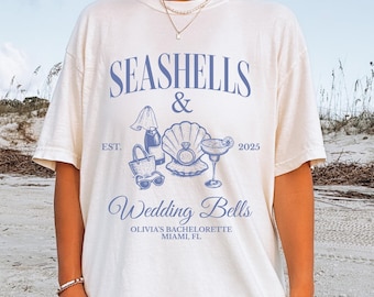 Op de afbeelding: Wit t-shirt met een blauw ontwerp. Het ontwerp bevat de tekst "SEASHELLS & Wedding Bells" en illustraties van schelpen, een cocktail en een strandtas. De tekst "OLIVIA'S BACHELORETTE MIAMI, FL" is ook opgenomen.
