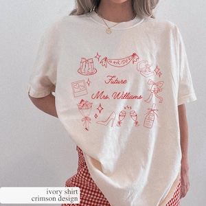 Può includere: T-shirt color avorio con un design cremisi con illustrazioni a tema matrimonio. Il design include uno striscione con la scritta "Bride", un portagioie, champagne e il testo "Future Mrs. Williams".