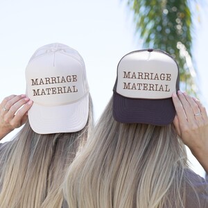 Puede incluir: Dos gorras de camionero con el texto "MARRIAGE MATERIAL" en letras marrones. Una gorra es de color crema con la parte trasera de malla blanca, y la otra es marrón oscuro con la parte trasera de malla negra. Las gorras son sostenidas por personas con pelo largo y rubio.