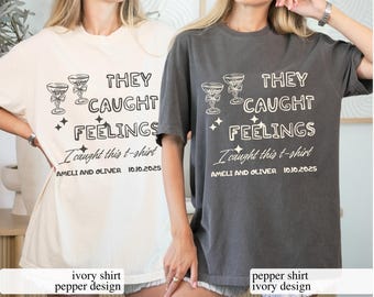 May include: Two t-shirts, one ivory and one pepper, each with the text "THEY CAUGHT FEELINGS" and "I caught this t-shirt". The design includes champagne glasses and the names "AMELI AND OLIVER" with the date "10.10.2025".