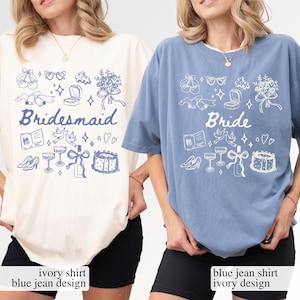 May include: Two t-shirts, one cream and one blue, with wedding-themed illustrations. The cream shirt says "Bridesmaid" in blue, while the blue shirt says "Bride" in white. Both shirts have matching graphic designs of wedding elements.
