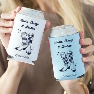 K&ouml;nnte beinhalten: Zwei Getr&auml;nke-K&uuml;hlmanschetten, eine wei&szlig;e und eine hellblaue, jeweils mit einem Design mit Cowboy-Stiefeln und dem Text "Boots, Booze & Besties" und "Nashville 2025".