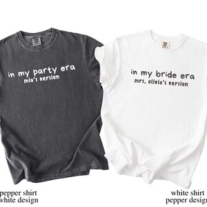 May include: Two t-shirts, one dark gray and one white, with text. The dark gray shirt says "in my party era mia's version." The white shirt says "in my bride era mrs. olivia's version."