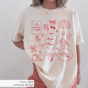 May include: Ivory-colored t-shirt with a crimson design featuring wedding-themed illustrations and text. The design includes phrases like "It's giving wifey," names "Oliver + Megan," and "future Mrs. Miller." The shirt also has the text "est. 2025."