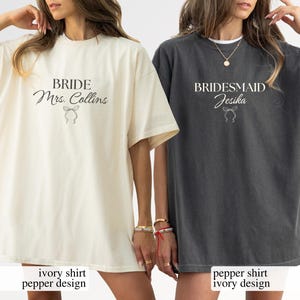 Peut inclure: Deux t-shirts amples, un ivoire et un poivre, sont présentés. Le t-shirt ivoire indique "BRIDE Mrs. Collins" avec un motif de nœud. Le t-shirt poivre dit "BRIDESMAID Jesika" avec un nœud similaire. Les deux t-shirts ont des manches courtes.