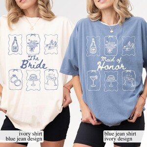 May include: Two t-shirts, one ivory and one blue jean, with matching designs. The ivory shirt says "The Bride" and the blue jean shirt says "Maid of Honor". Each shirt features blue illustrations of a bottle, flowers, and shoes.