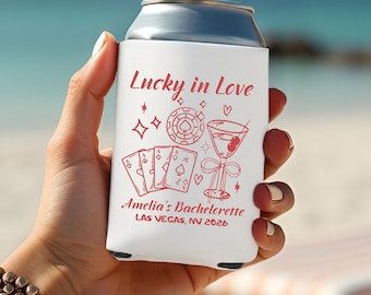 May include: A white can cooler with a red graphic design that includes the text "Lucky in Love", playing cards, a casino chip, a martini, and the text "Amelia's Bachelorette LAS VEGAS, NV 2026".