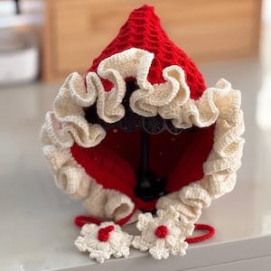 May include: A red and cream-colored crochet bonnet with a ruffled brim and floral accents. The bonnet is displayed on a black stand, showcasing its intricate design and texture. The bonnet has a textured red crown and cream-colored ruffles.